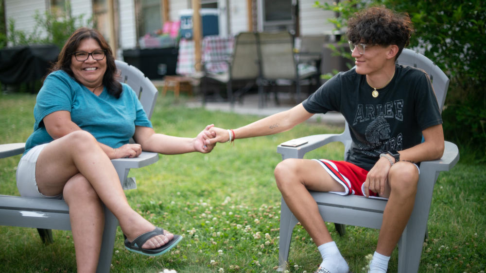A teenager and his mom sit smiling and holding hands on lawn chairs in a front yard.