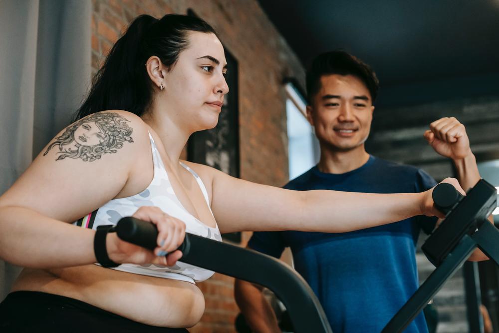 An overweight woman does cardio exercise on an elliptical while a physical fitness trainer encourages her.