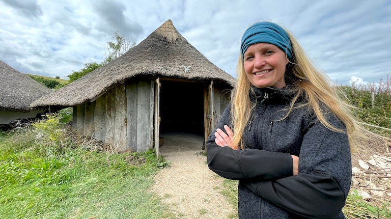 A woman smiles outdoors in front of a hut.