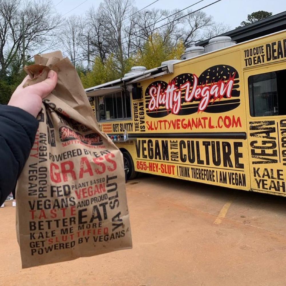 An outstretched arm holds a paper bag of food in front of the Slutty Vegan food truck in Atlanta.