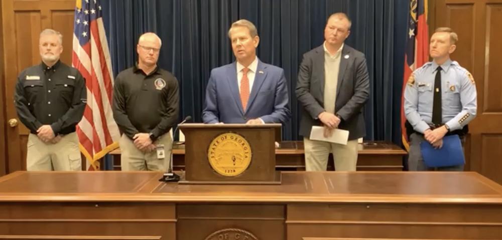 Governor Brian Kemp is shown standing behind a desk and lectern flanked by state officials.