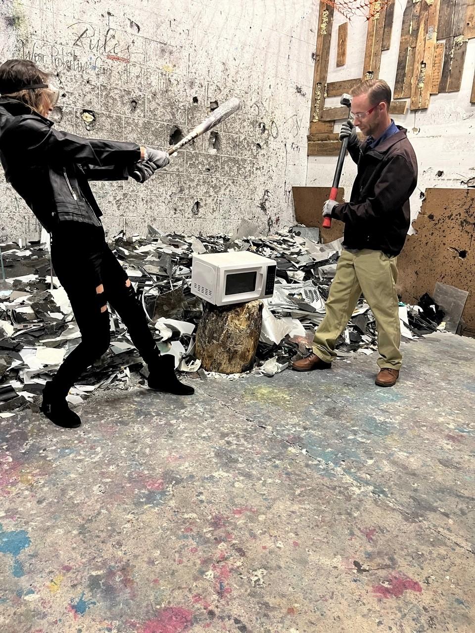 Two people, one wielding a bat and the other a sledgehammer, are poised to smash a small microwave in a cinderblock room.