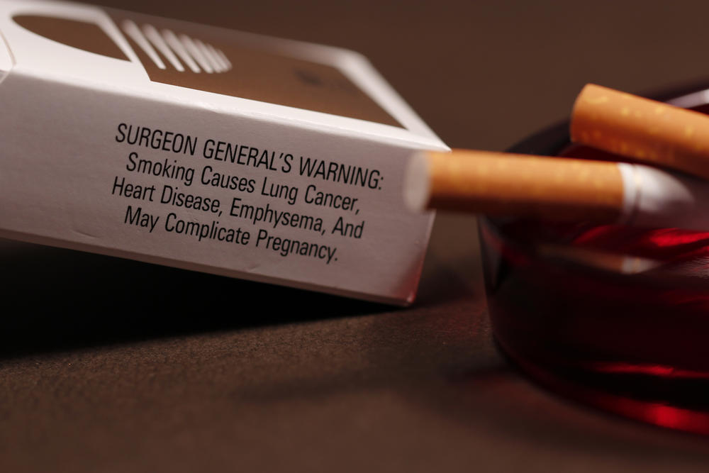In its background, this image depicts an opened pack of cigarettes with its side-panel health warning to would-be smokers stating some of the ill effects attributed to smoking, and in the foreground, a ruby-colored glass ashtray containing the butts of two cigarettes. 