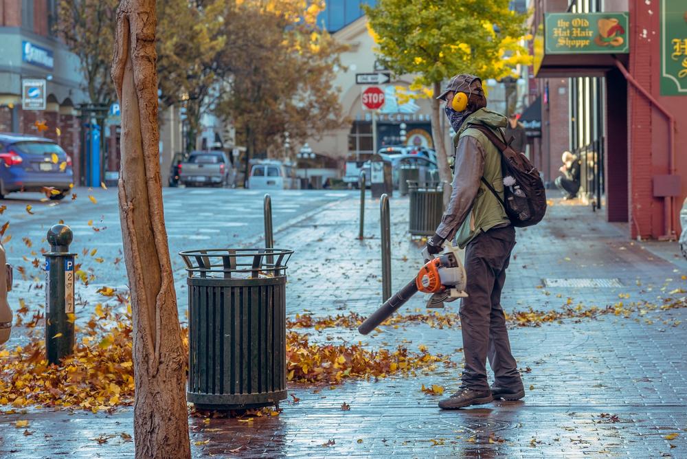 A man uses a leaf blower on a city sidewalk