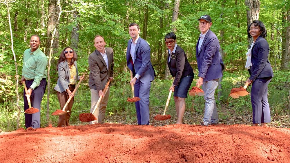 Seven people are shown holding shovels in a groundbreaking ceremony in a wooded environment.