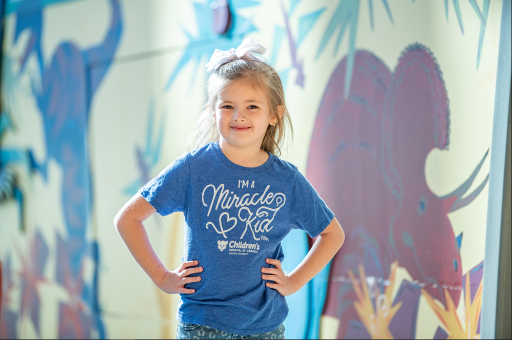 Reagan Milford stands with hands on hips with a blue shirt that says "I'm a miracle kid."