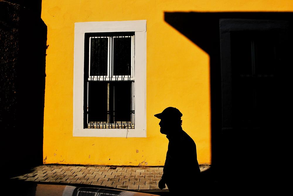 Silhouette of a man walking past a yellow building with a white framed window.