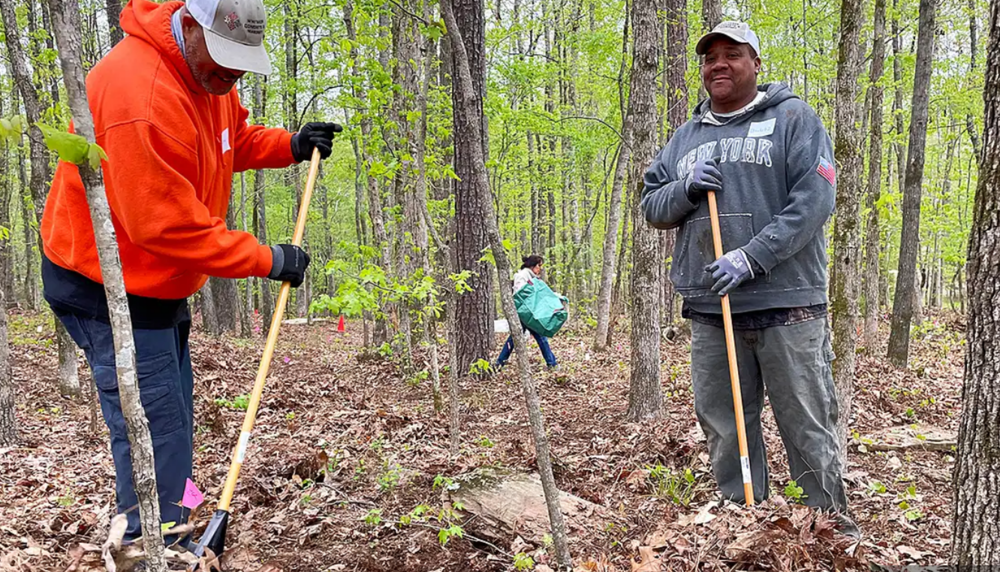 Two men hold rakes in a forested scene.