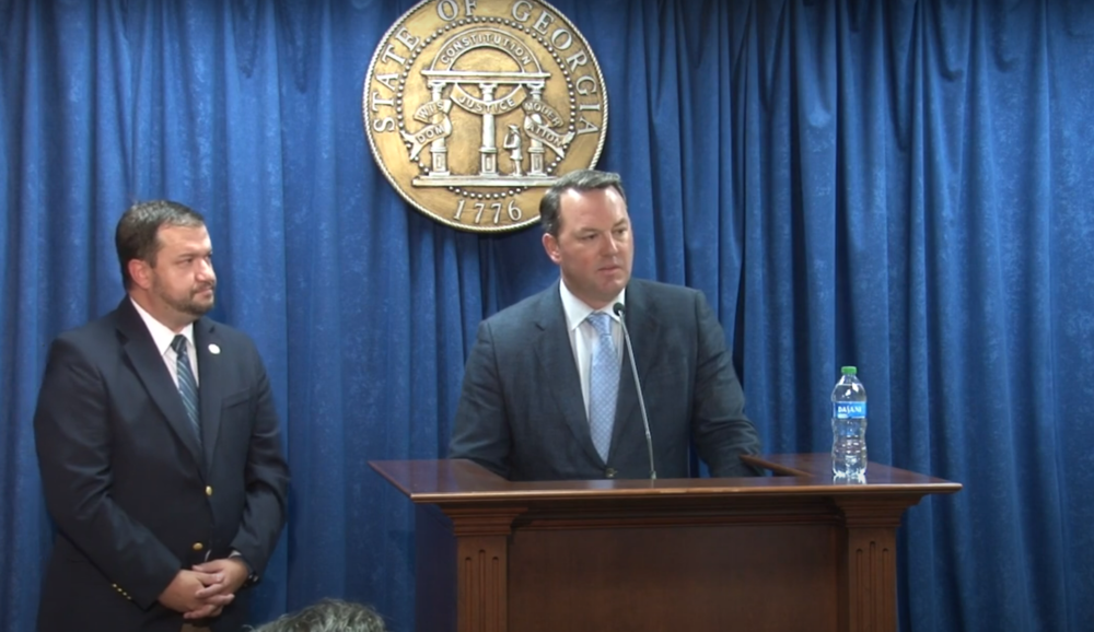 Burt Jones is seen speaking at a podium in front of a plain curtain backdrop and an official Georgia seal. Dallas State Senator Jason Anavitarte is shown standing to his left.