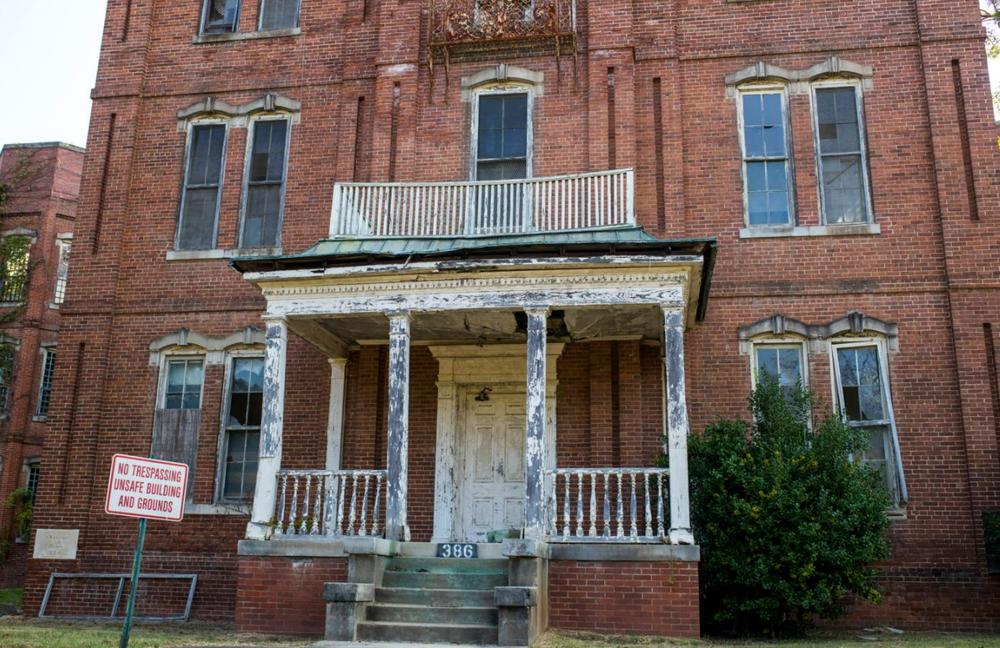 The run-down facade of an old brick building is shown with a sign in front of it that says "No Trespassing Unsafe Building and Grounds."