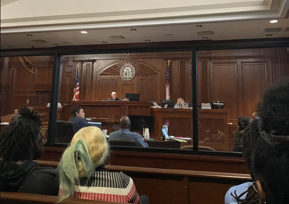 Public defender Tobe Karrh (left in suit) looks on at evidence projected onto a screen in the felony murder trial of his client Varshan Brown (center in blue shirt) on Sept. 12, 2023, in the killing of his cousin Latoya James. James was killed by police during a drug raid, according to court testimony.