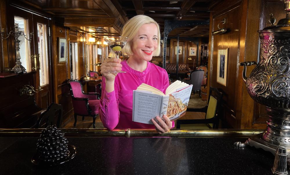 Lucy Worsley sitting at a bar with a cocktail in one hand and an open book in the other.