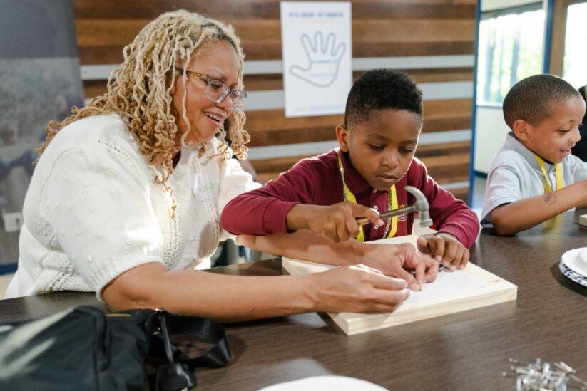 A woman helps a young boy hammer a nail into a piece of wood while seated at a table