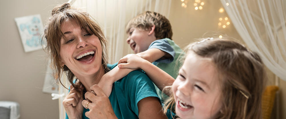 A woman in scrubs with a young boy on her back and a young girl is smiling