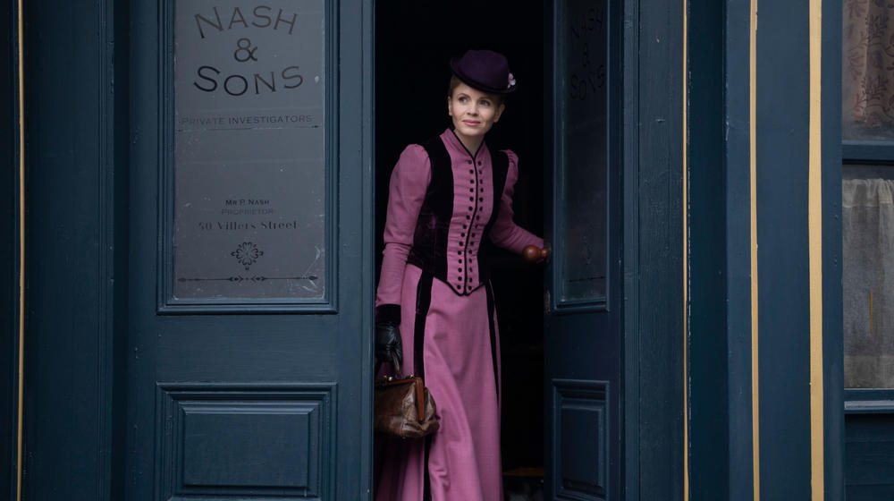 A woman in Victorian period dress stepping out of a building.