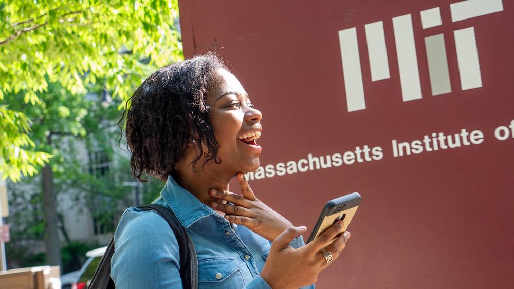 A young woman holding a cell phone and laughing in front of a sign for MIT.