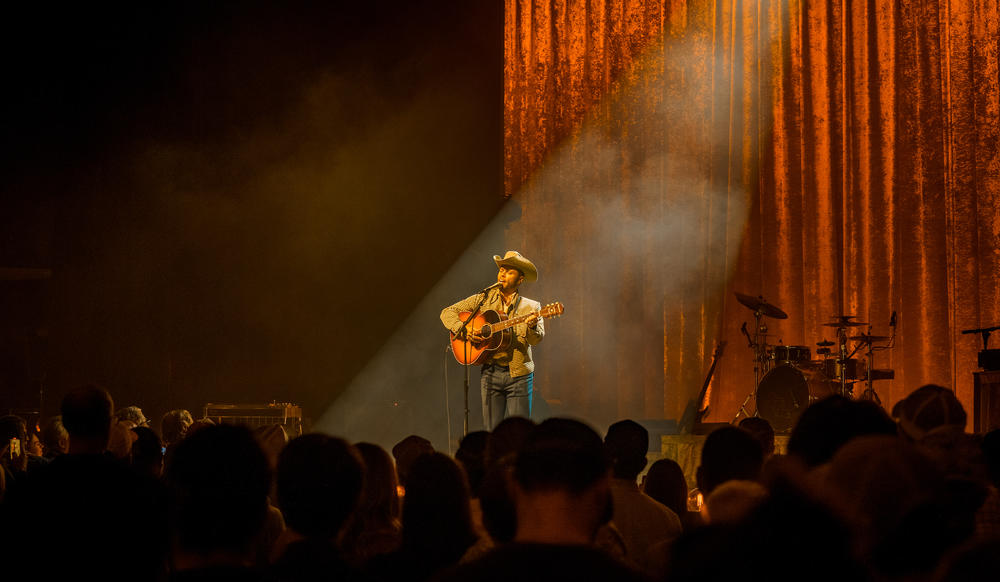 A man in a cowboy hat holding a guitar on a stage with a spotlight on him.