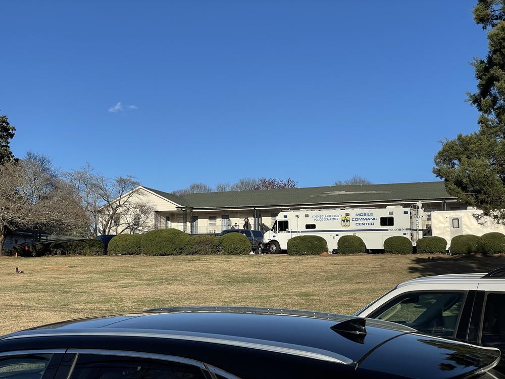 A police mobile command center vehicle is shown in the distance across a grass lawn and in front of apartment units.
