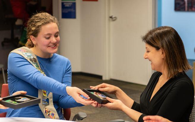 Lily Crick hands Dr. Mandy Cohen a framed public health fun patch created by Troop 14567.