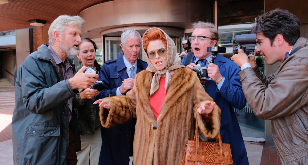 A woman in 60s period dress surrounded by photographers and reporters.