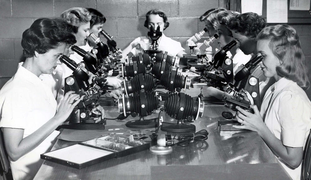 A black and white image of a group of women at a table looking into microscopes.