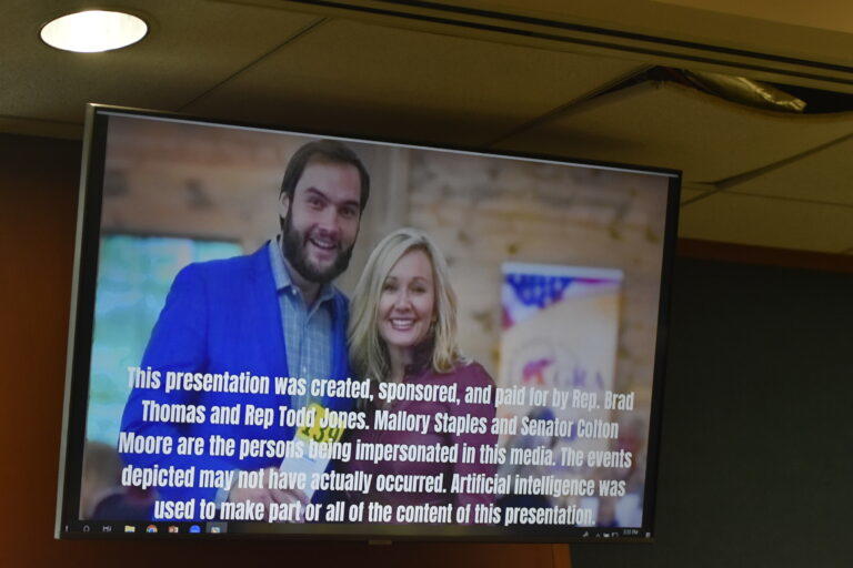 TV in a legislative committee room showing an AI image of Sen. Colton Moore and Freedom Caucus State Chair Mallory Staples
