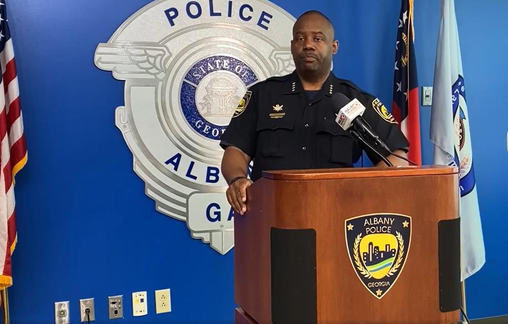 Albany Police Chief Michael Persley is shown standing at a podium, indoors and flanked by three flags.