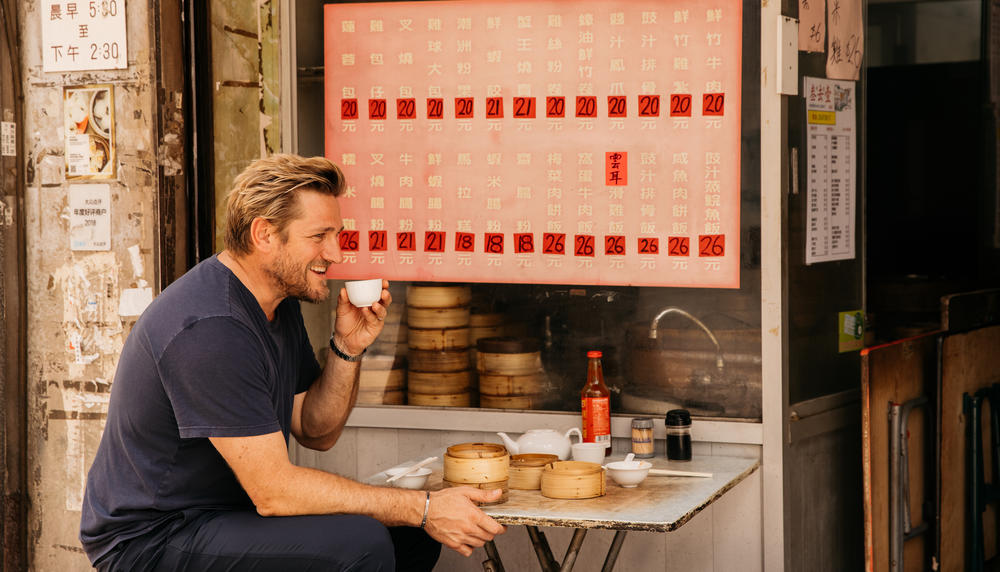 A blonde man drinking tea from a small cup at an outdoor cafe table in Hong Kong