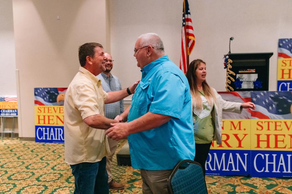 Two people are shown shaking hands inside a building with an American flag and signs saying "Steven Henry Chairman."