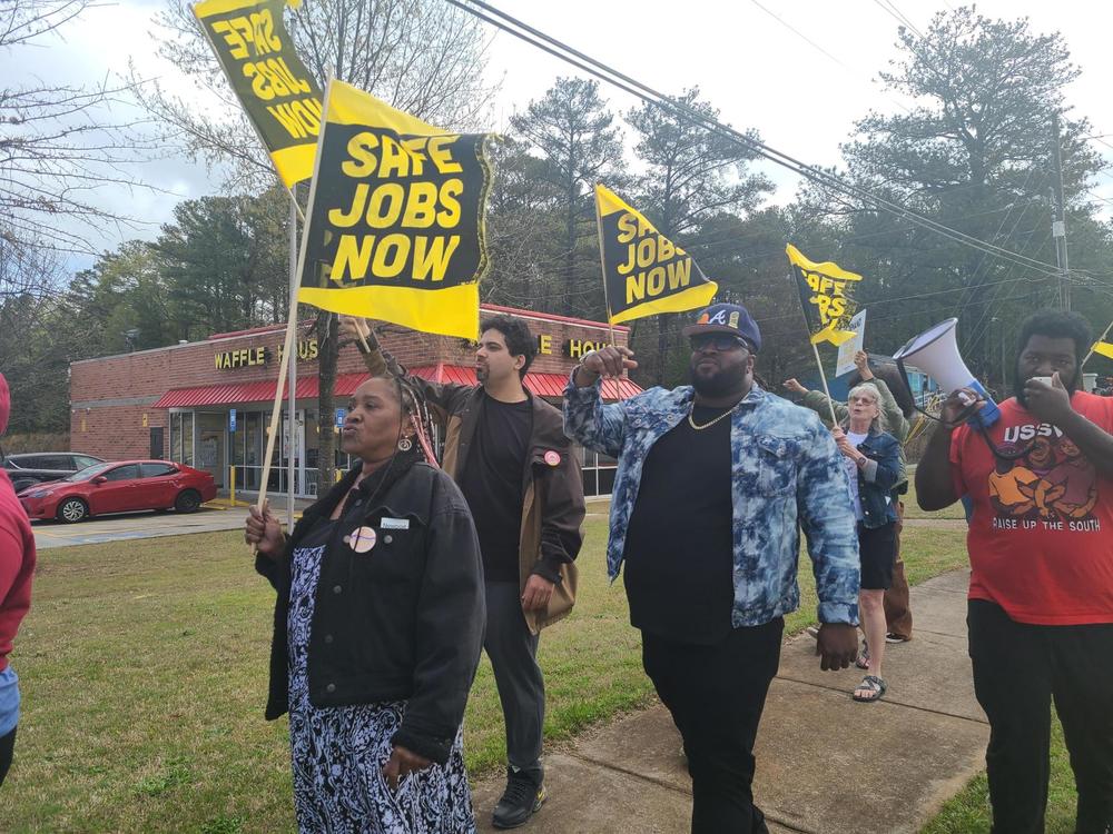 People walk down the sidewalk holding yellow and Black flags that say "Safe Jobs Now". A waffle House can be seen in the back