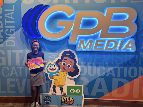 Jewel Rich stands holding her art beside a "Lyla in the Loop" standee.