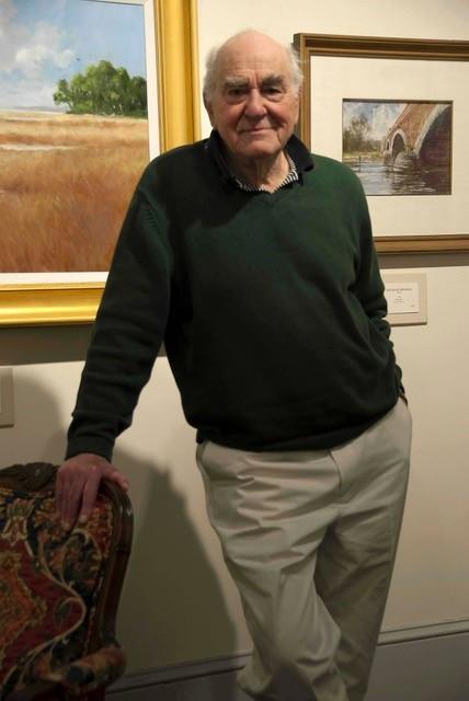Ray Ellis is shown standing in front of two of his paintings and leaning against a chair in this posed photograph.