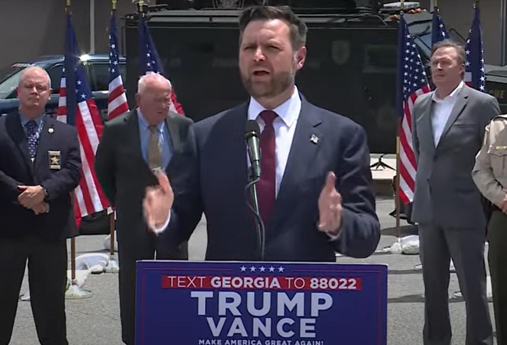 Ohio Sen. J.D. Vance is shown speaking from behind a podium. He is flanked by other men and flags.