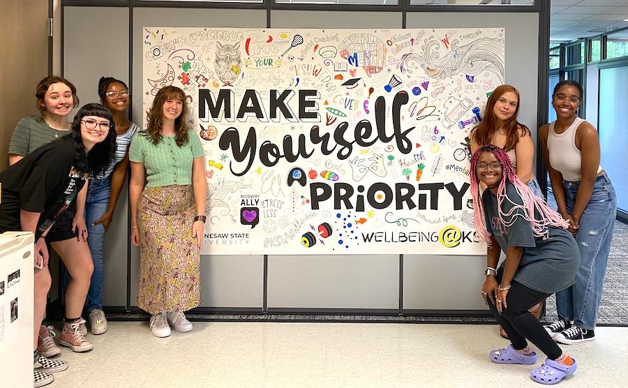 A group of students at Kennesaw State University stand with a sign that says "make yourself a priority"