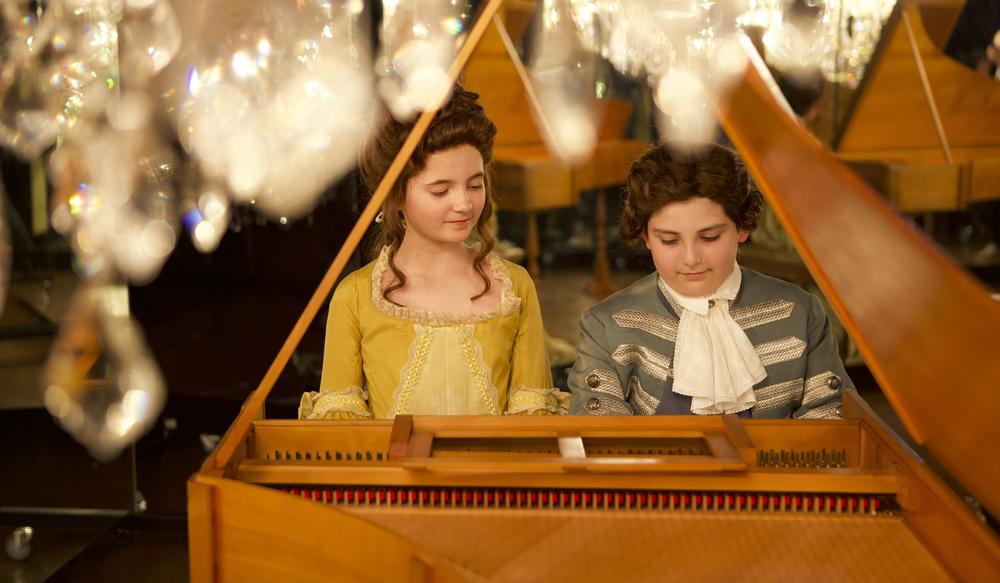 Two children in period costumes sitting at a piano.