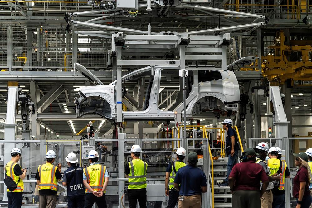The unfinished frame of a sports utility vehicle hangs above a group of about a dozen people wearing safety vests and hard hats inside a factory.