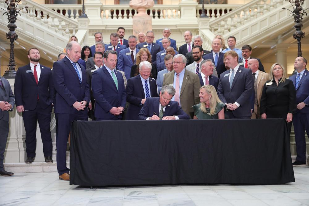 Governor Brian Kemp is shown signing the Amended Fiscal Year 2025 Budget, seated at a table, with dozens of officials standing behind him.
