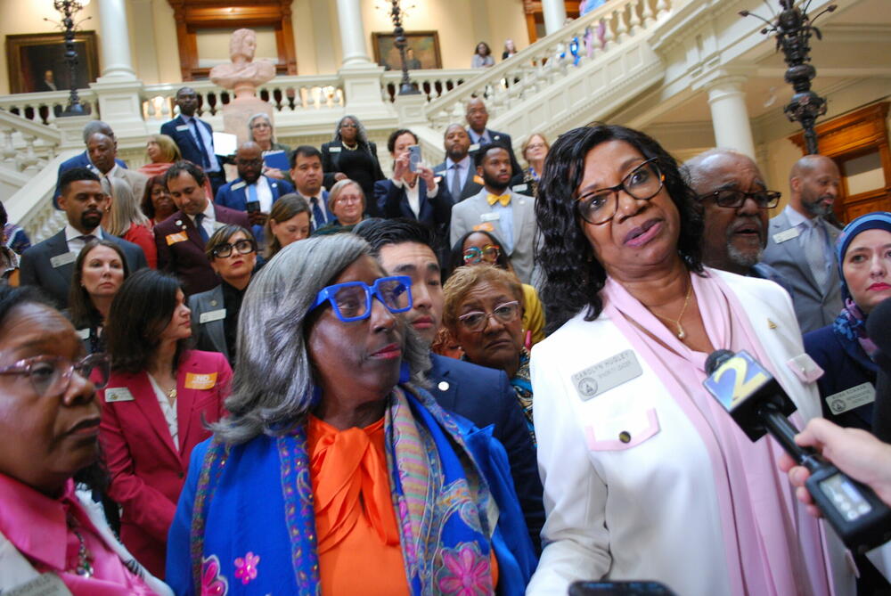 Democratic members of the Georgia House are shown standing on the steps inside the Georgia capitol, with one member speaking into a reporter's microphone.