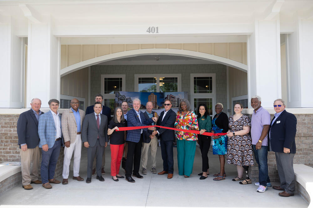 A group of at least a dozen people are seen holding a ribbon about to be cut in front of a building.