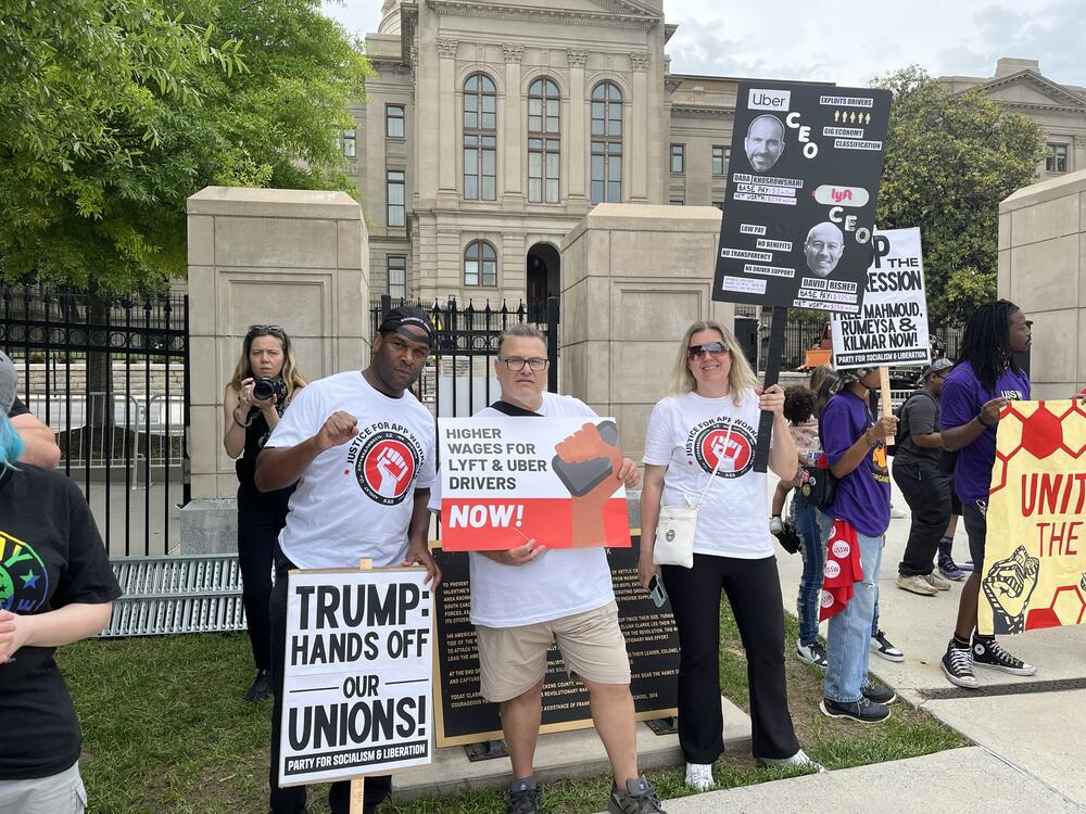 Three people in white shirts that say justice for app workers holding pro union signs.