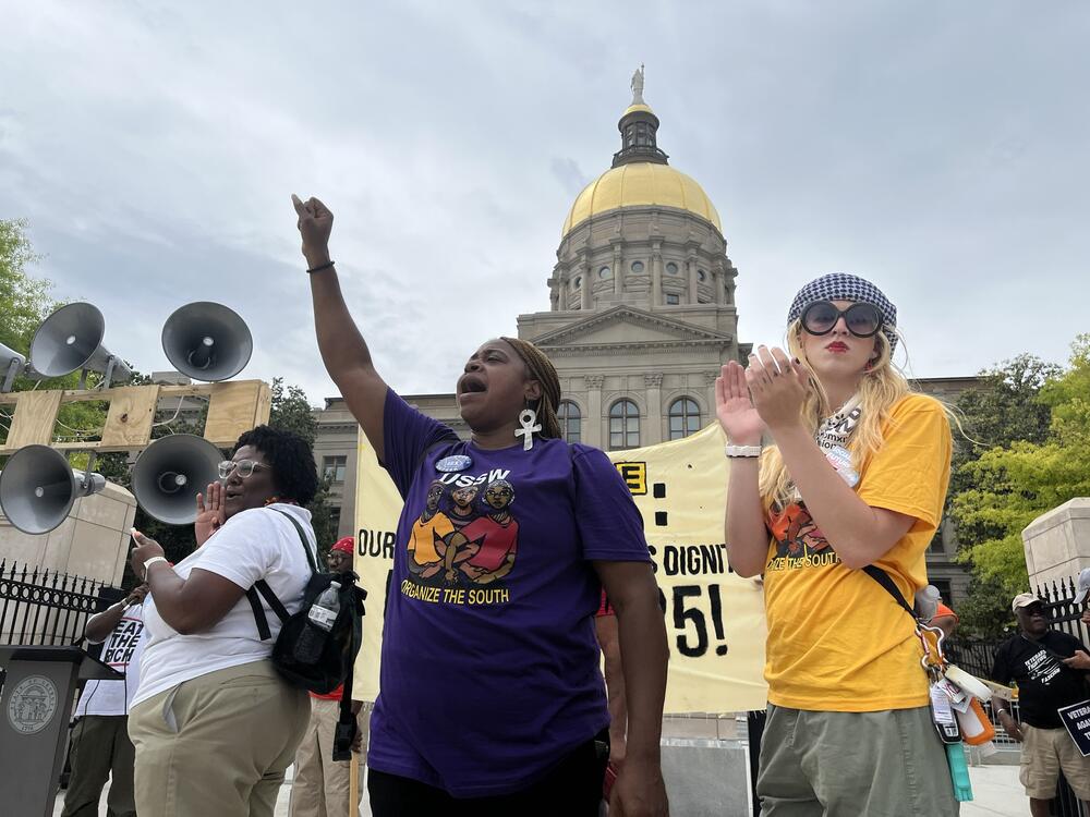 Three women in Union shirts stand in front of the state capitol, one has her first up.