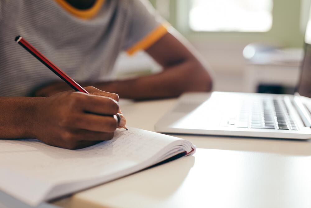 An elementary school aged child writes in a book next to laptop computer