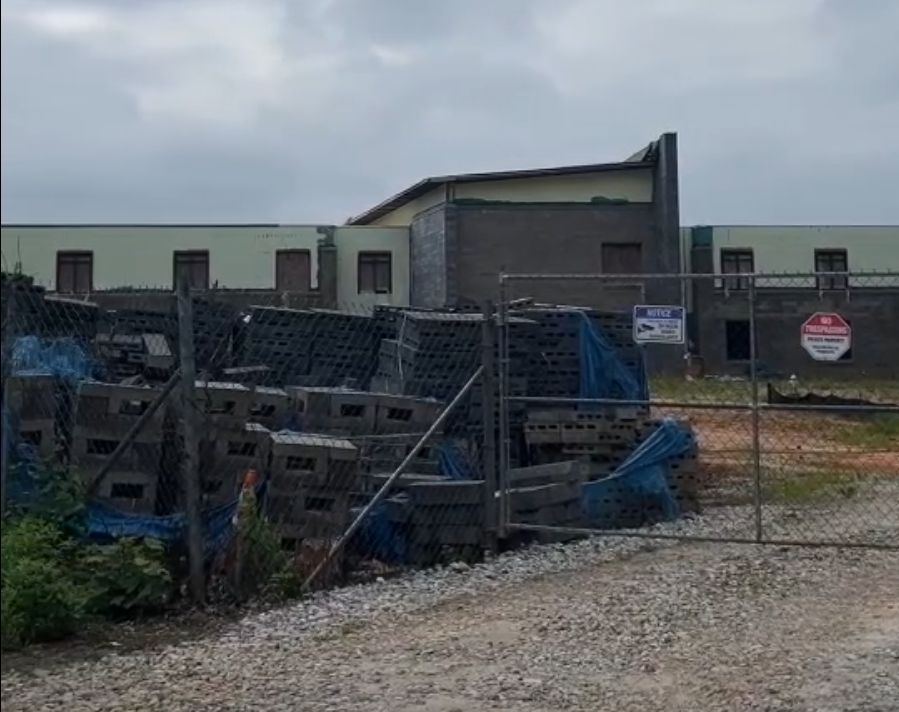 a fence with concrete blocks and other building materials in front of a large partially built building with boarded windows