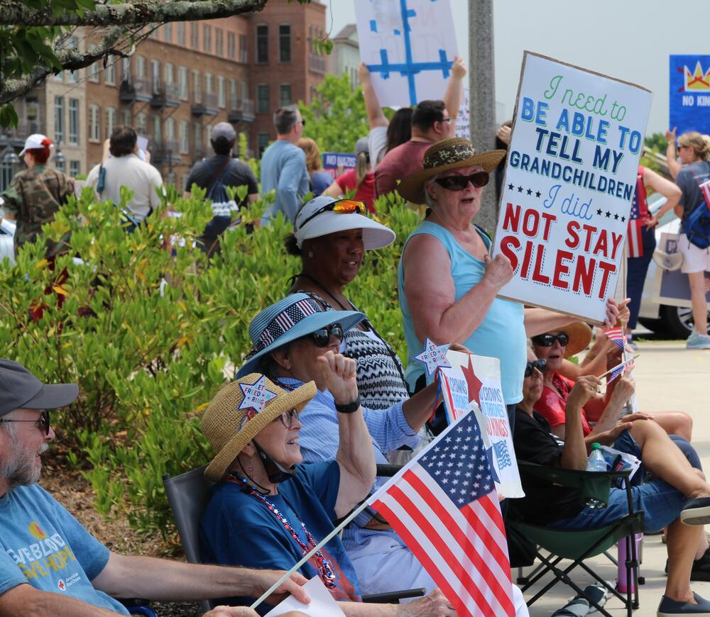 Older adults gather on the sidewalk in Downtown Woodstock June 14, 2025, waving flags and signs. 