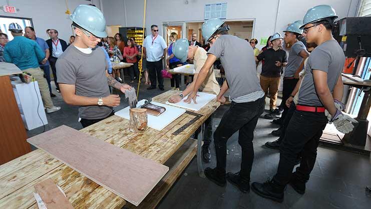 A group of young people wearing hard hats are seen in a wood working shop.
