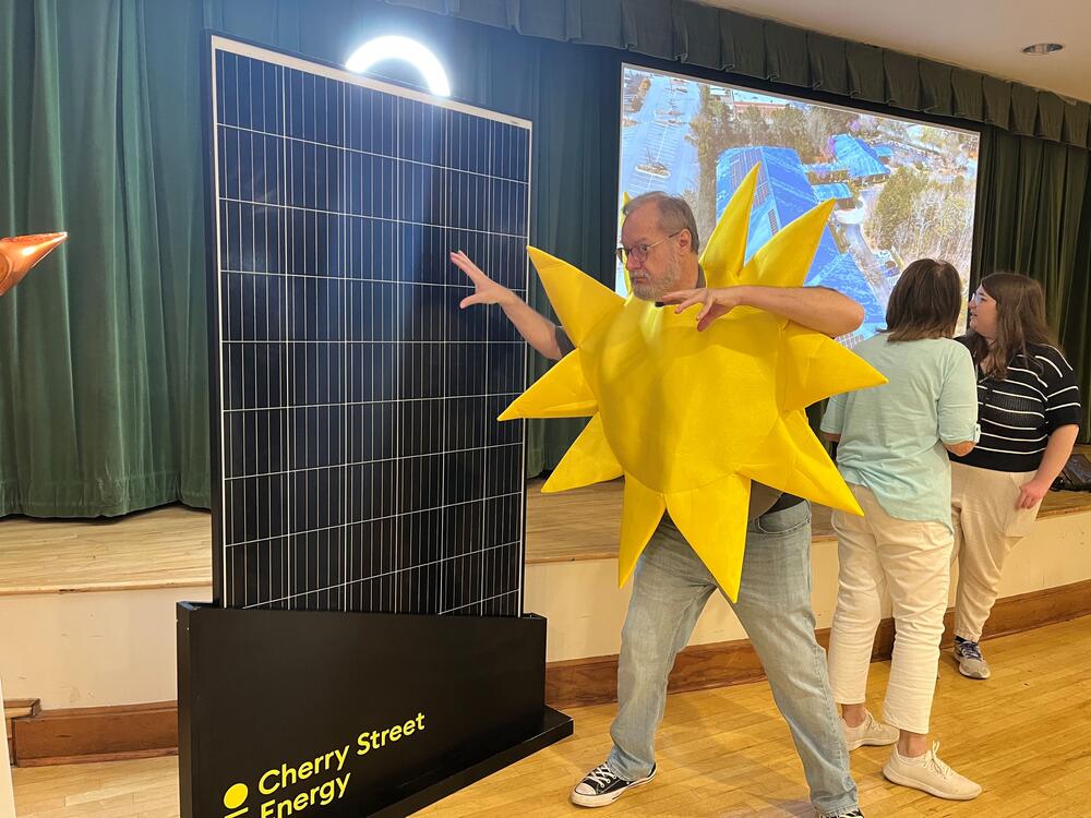 A man dressed in a sun costume points at a solar panel as though he's powering it.