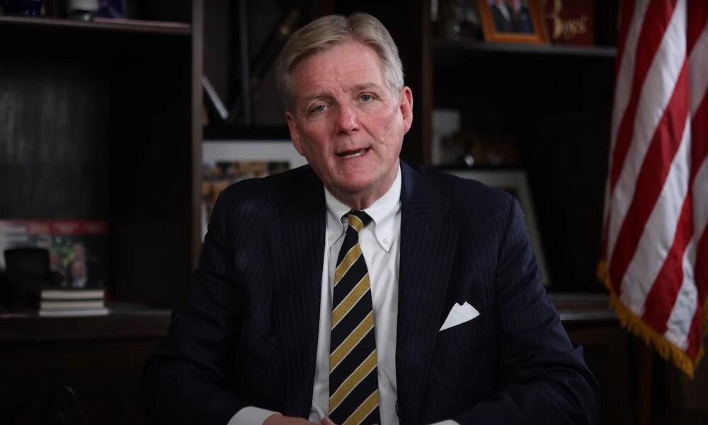Brant Frost IV is shown seated and speaking to the camera in his office with an American flag in the background.
