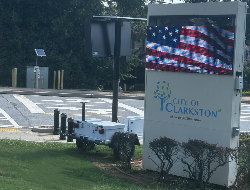 An LED screen shows a waving American flag on a welcome sign for the City of Clarkston. The text beneath reads "Where possibilities grow"