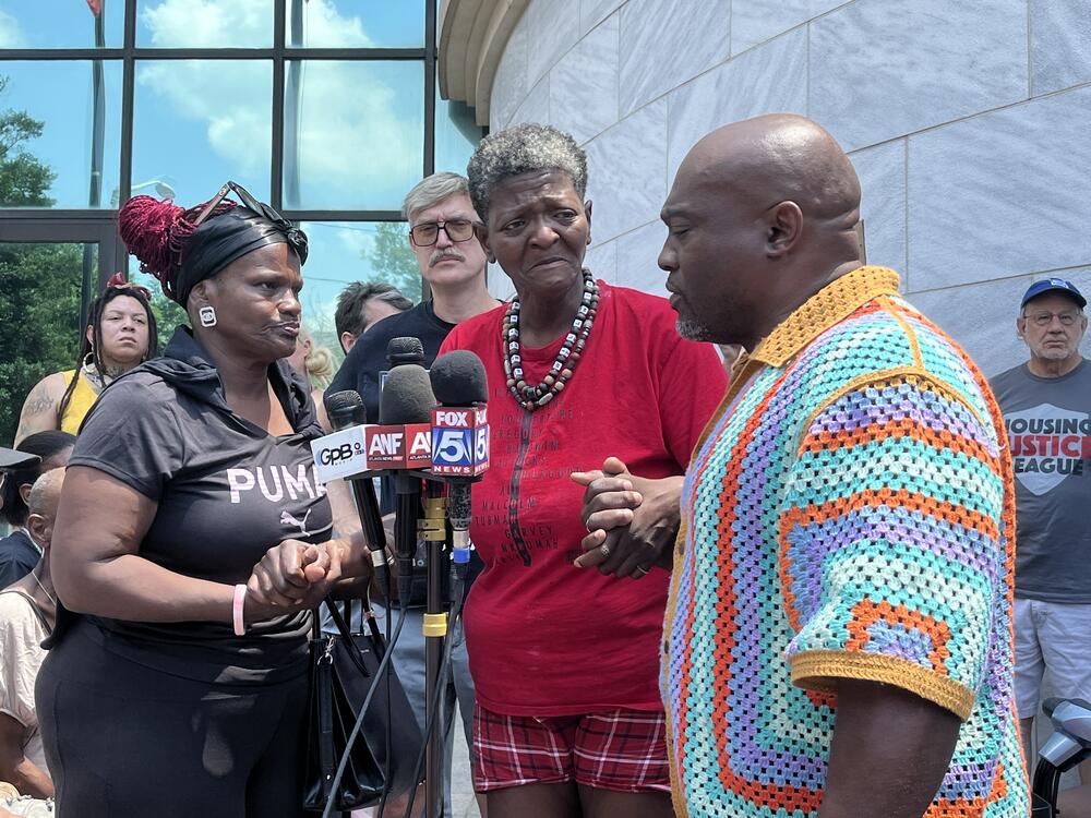 A woman stand in front of microphones with someone holding her hand on both sides. She is crying.