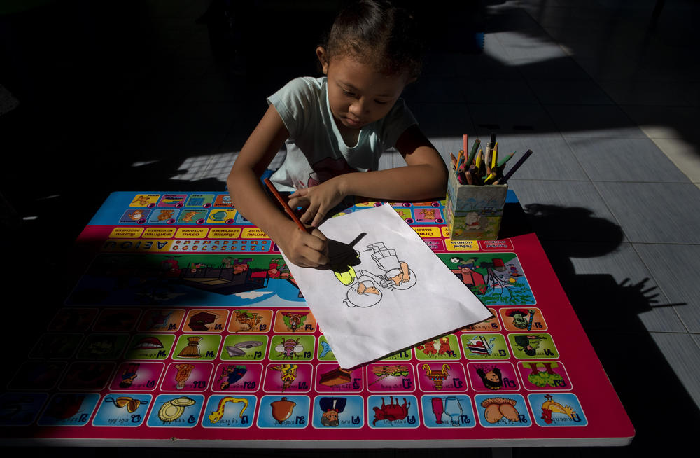 Warisara Klawkla, a daughter of teacher Watcharee Klawkla, colors a drawing made to educate children on COVID-19 prevention methods while her mother help cooking a meal at Makkasan preschool in Bangkok, Thailand, Wednesday, June 24, 2020.